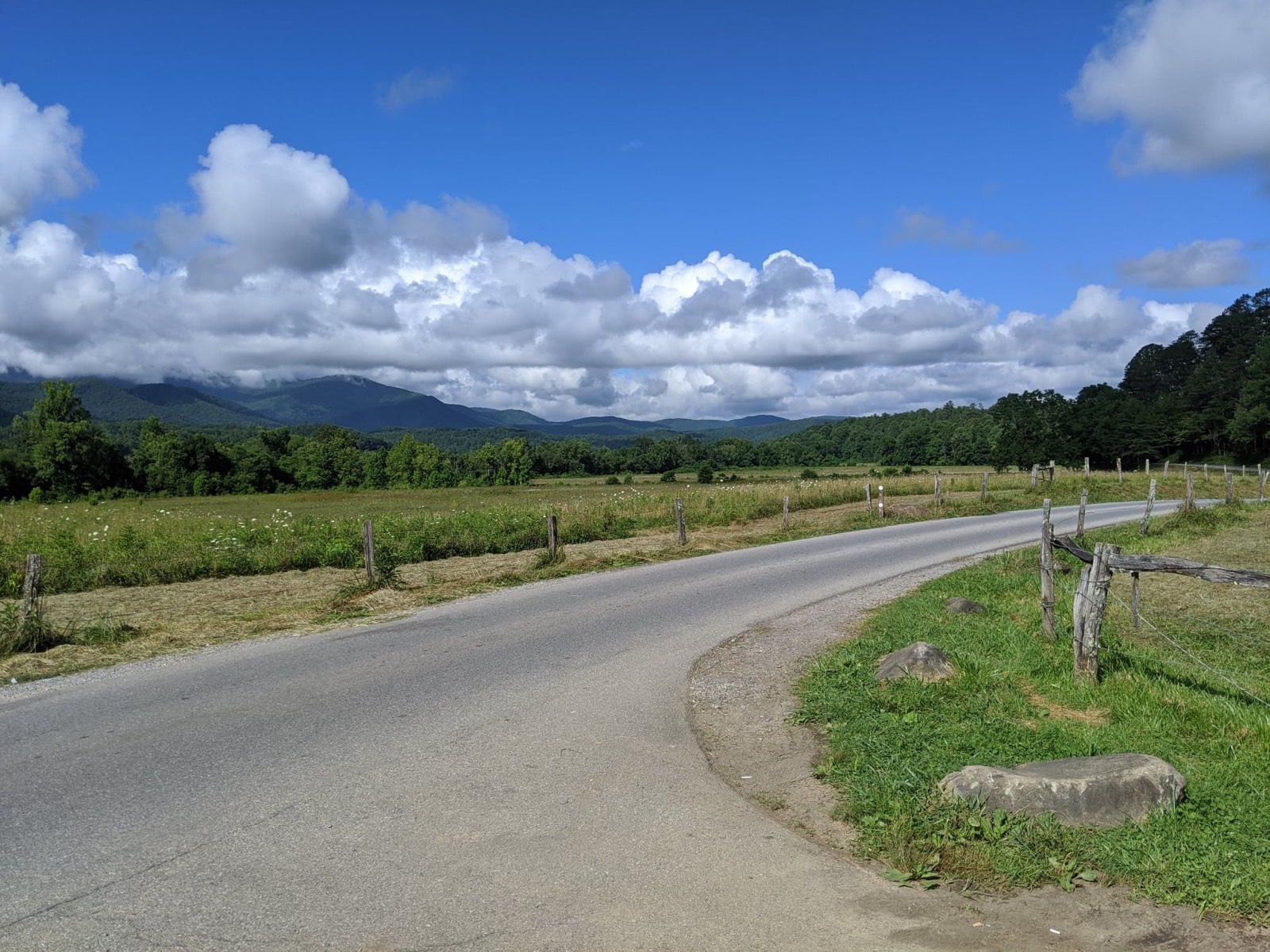Cades Cove Loop Road curving past split-rail fence and open valley fields, with the Great Smoky Mountains under summer cloud in the distance — NPS photo near the John Oliver Cabin
